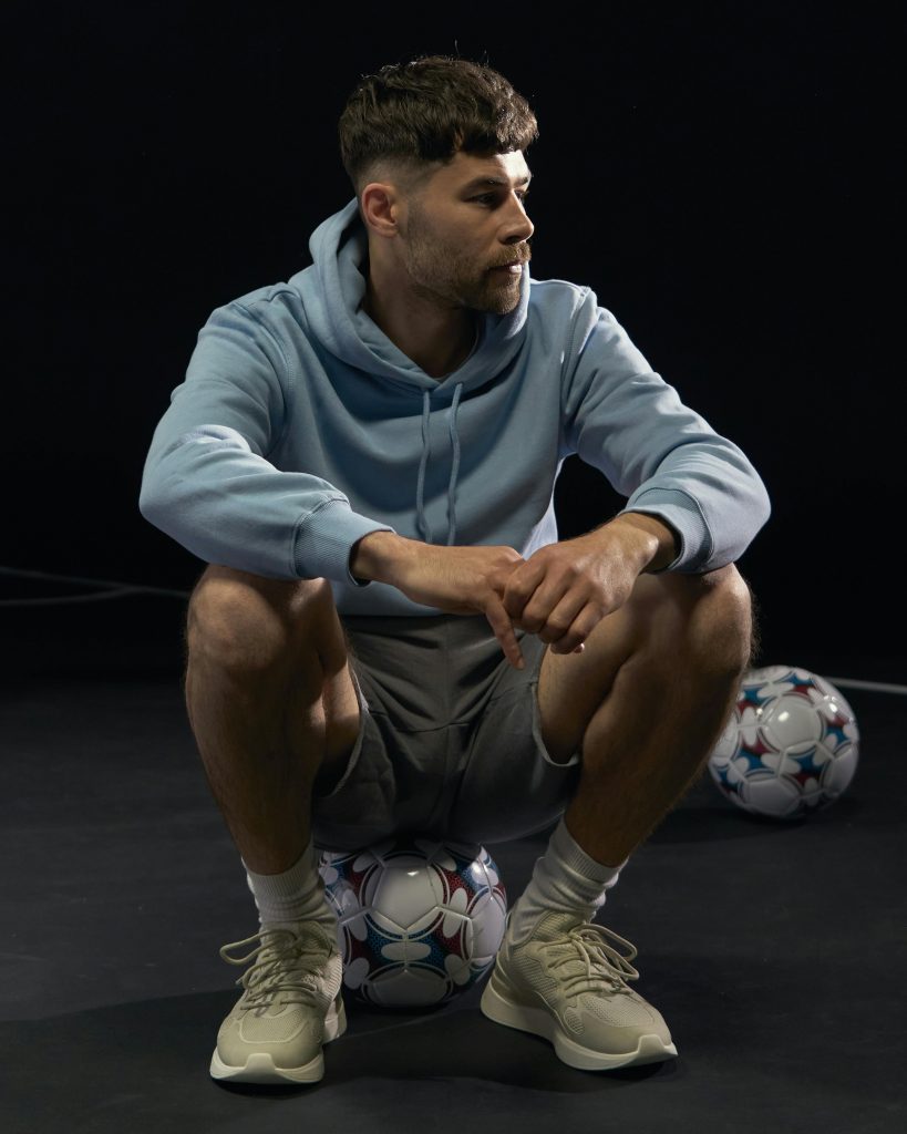 Young man in hoodie sitting on soccer ball in studio setting. Sporty and relaxed vibe.