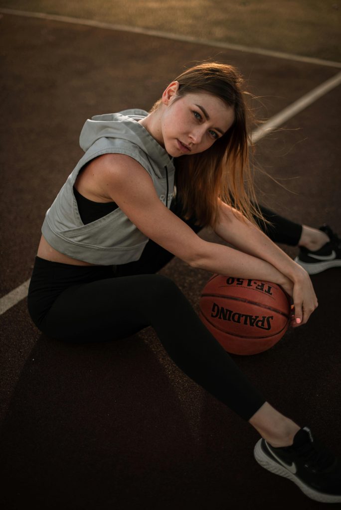 Stylish young woman sitting on basketball court with ball, showcasing athletic fashion and attitude.