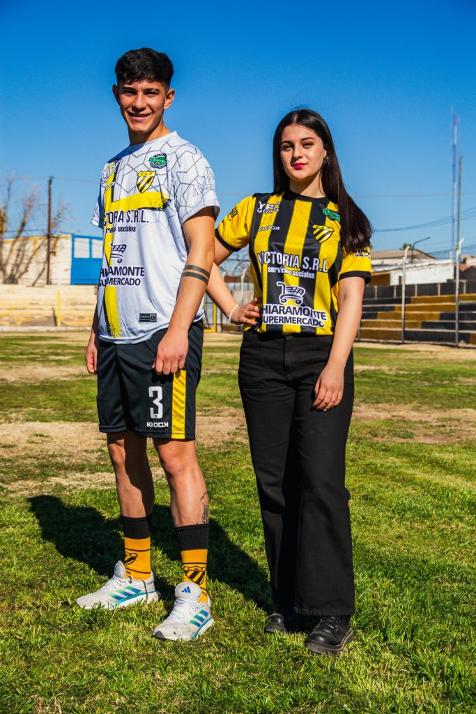 A teenage soccer player and companion in team uniforms stand on a sunny outdoor field.