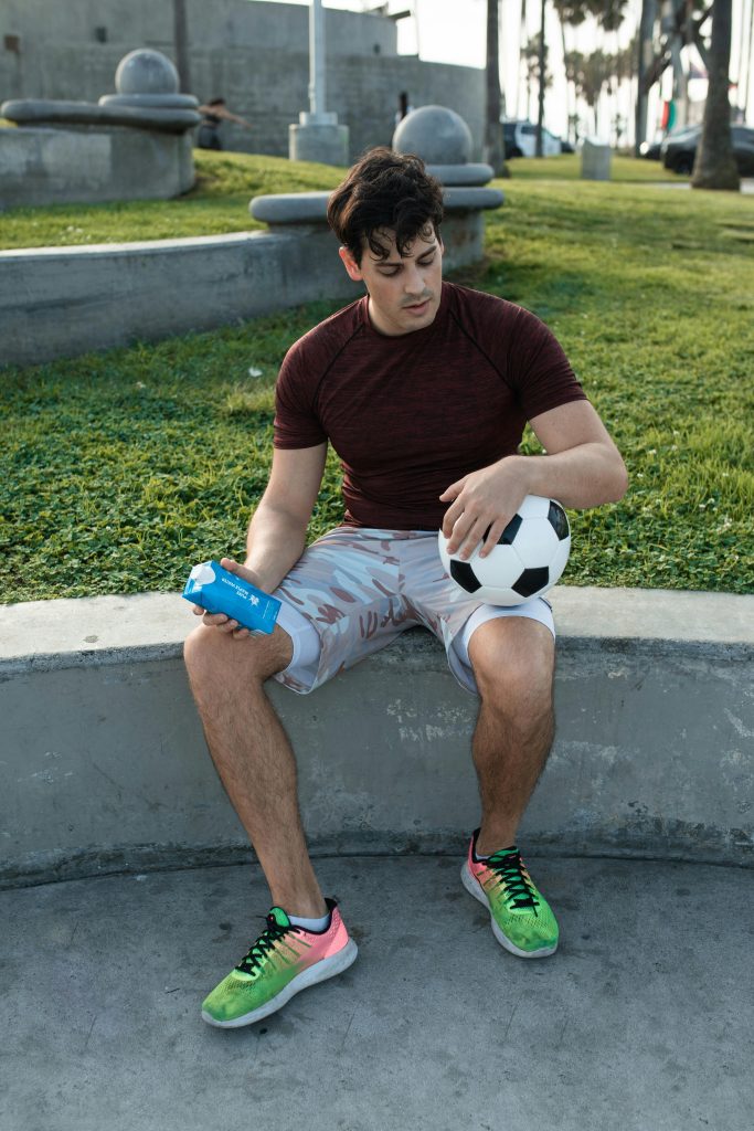 A young man sitting outdoors with a soccer ball and water bottle, enjoying a sunny day.