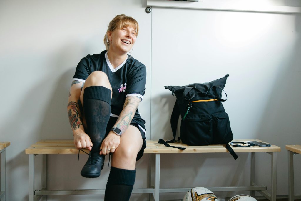A female soccer player laughing while putting on boots in a locker room.